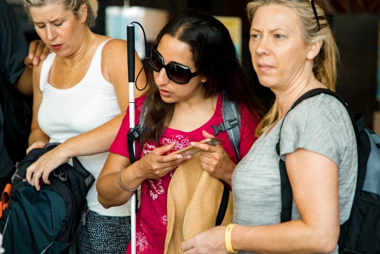 A blind woman with dark glasses and a walking stick tilts her head to hear her smartphone.