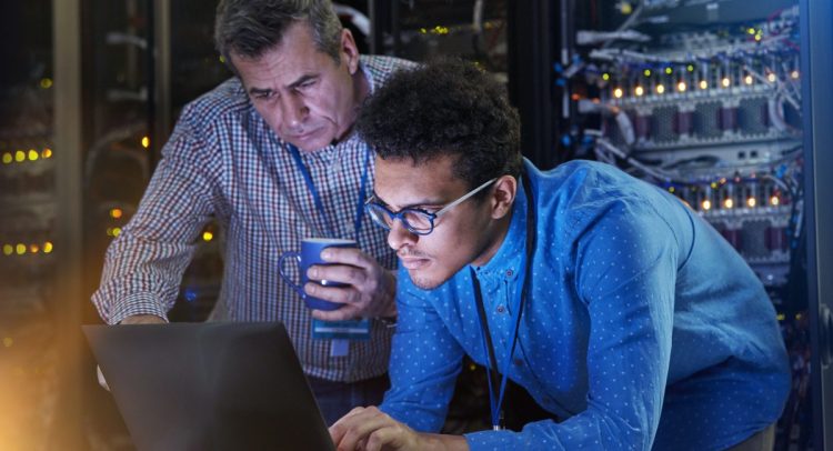 Focused male IT technicians working at laptop in dark server room