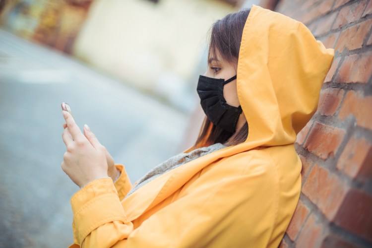 A young woman stares at her phone while wearing a face mask in a polluted city