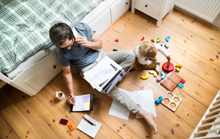 Father working from bedroom floor with his child playing nearby
