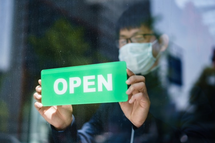 Business owner hangs open sign on door while wearing protective mask