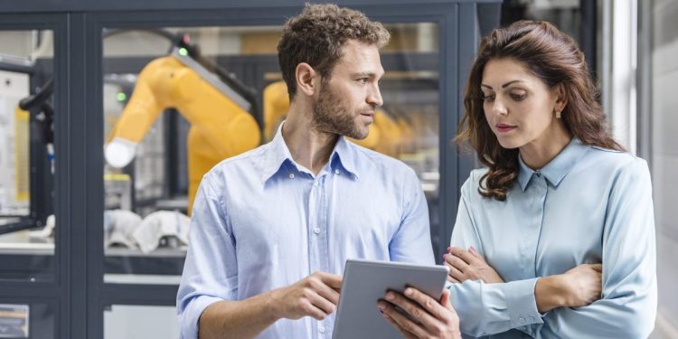 Two colleagues discuss findings on a tablet with advanced robotics technology in the background.