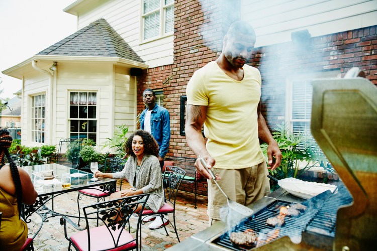 Man barbecues meat on the grill while two women and a man wait to eat
