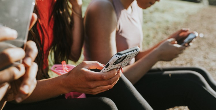 Three woman sit outside in the sun and hold their smart phones. They all look at their screens, while using fingers and thumbs to type. Conceptual with space for copy.