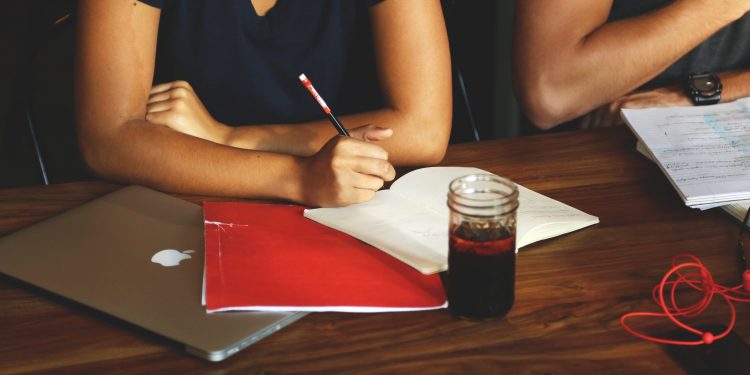 Table in front of a person in a meeting. Person is writing in a notebook, with a laptop, drink, and folder next to them.