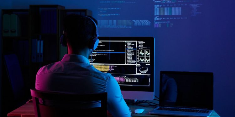 Software developer writing code on a desktop computer with a laptop next to it. Code is also projected on the wall. Developer appears to be working from home at a desk wearing headphones