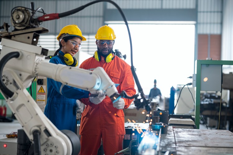 diversity in robotics showing female and male in working safety gear speaking collaboratively next to a robot arm also pictured
