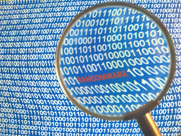 Magnifying glass over the word RANSOMWARE in red letters. Background of binary code on a computer screen.