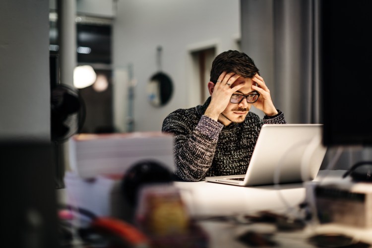 Young casual businessman wearing glasses is sitting in front of his notebook holding his head in concentration.