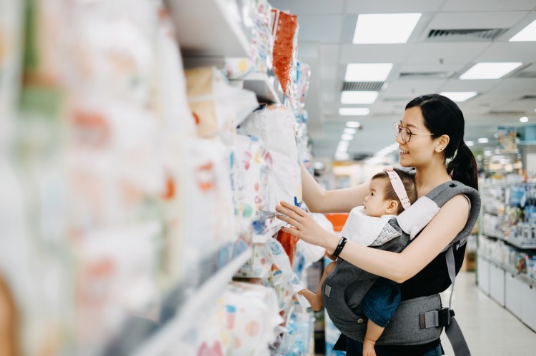 Young Asian mother carrying cute baby girl shopping for baby product in a shopping mall and is looking at a variety of diapers