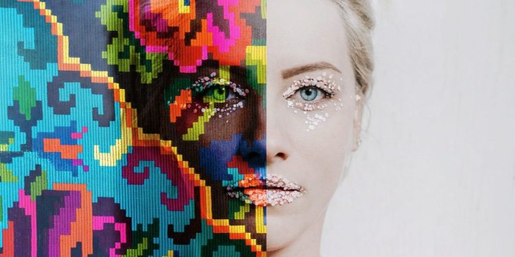Image of woman against a blank wall. The left side of her face is covered in colorful makeup, whereas the right side of her face is covered in pale, monochromatic makeup.