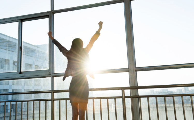 Employee value concept: Businesswoman looking through the window with arms raised.