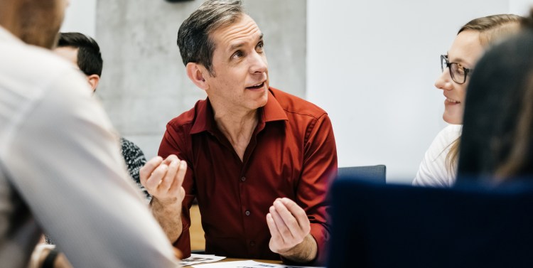 A caucasian man is speaking in a business meeting surrounded by colleagues listening to him. He uses his hands and seems convinced and focused.