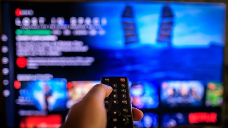 TV remote control in the foreground, Video on demand screen in the blurry background, streaming Netflix