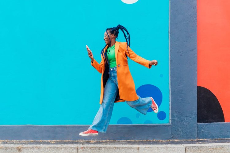Woman with braids looking at online fashion on a cell phone against a blue wall.
