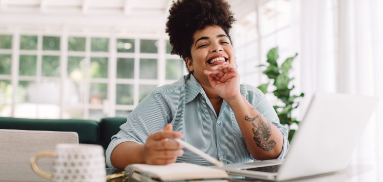Wide angle shot of happy female sitting at desk looking at camera. Attractive woman smiling while working from home.