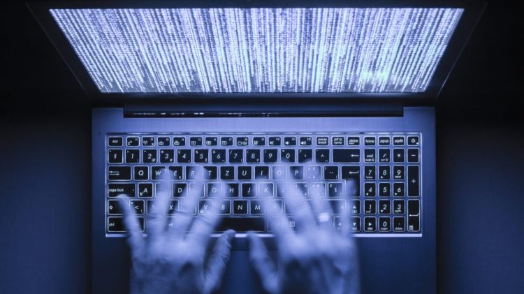 Blurred hands are typing on a laptop computer in the dark with illuminated keyboard and illegible mystic program code visible on the screen.
