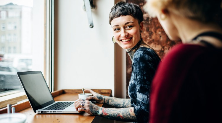 A young tattooed short-haired woman sitting in a bright café. She faces towards someone and smiles while a laptop is in front of her on the table.