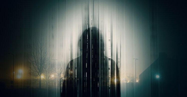A double exposure of a Silhouette of a mysterious hooded figure without a face, in a city at night.