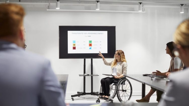 Disabled businesswoman giving presentation to colleagues. Female executive sitting on wheelchair while discussing with team. They are in meeting at workplace.