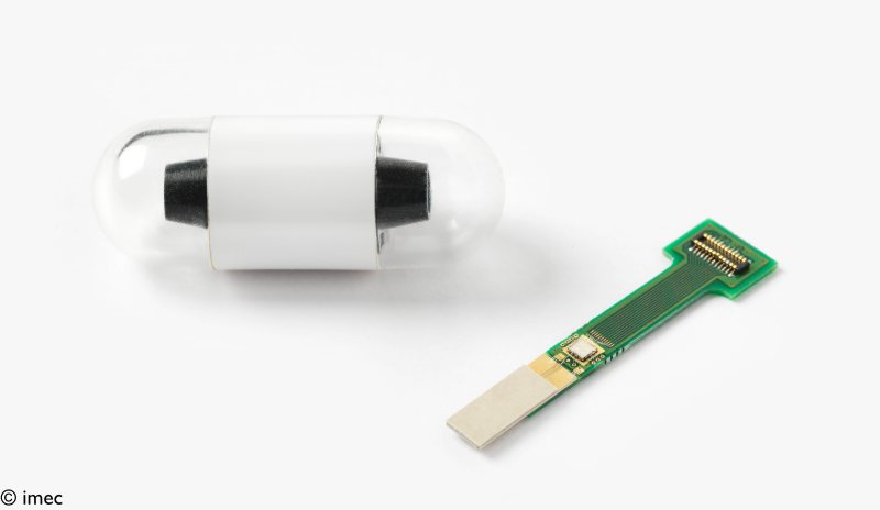 clear capsule containing a tubular device with black bumpers on either end, next to a flat, t-shaped circuit board.