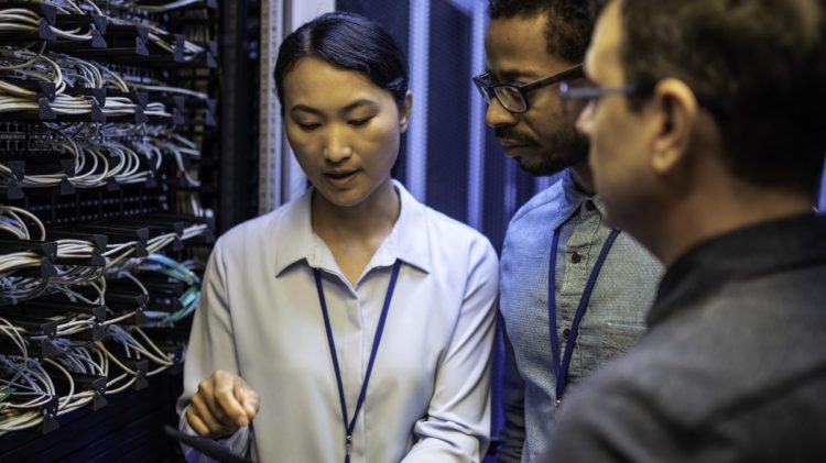 Three IT technician looking at a digital tablet and talking while standing next to a server in a data center.