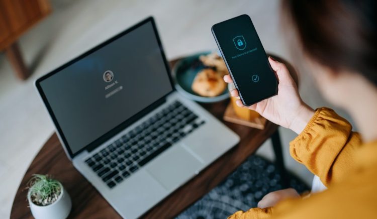 Young Asian woman logging in to her laptop and holding smartphone on hand with a security key lock icon on the screen, sitting in the living room at cozy home. Privacy protection, internet and mobile security concept