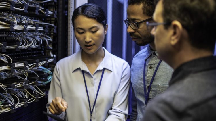 Three IT technician looking at a digital tablet and talking while standing next to a server in a data center.