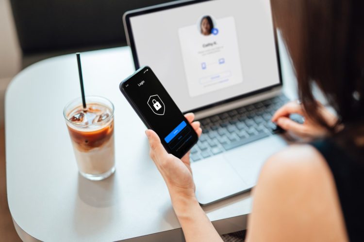 Young Business Woman Logging In Online Security System On Laptop With Mobile App On Smartphone