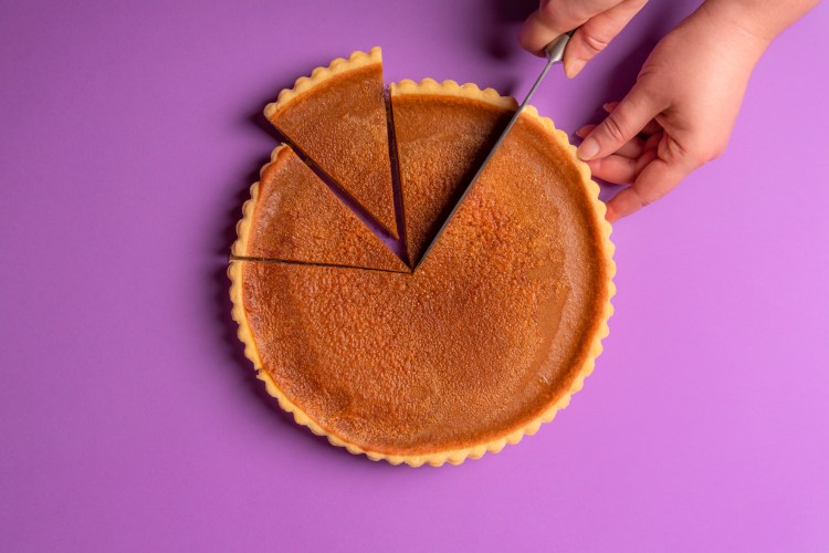 Woman hands slicing a pumpkin pie on a purple background. Minimalist culinary concept. Homemade sweet pie. Traditional holidays dessert. Top view.