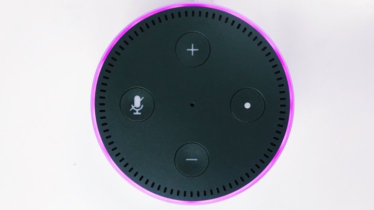 A close-up top view of a black Amazon Echo Dot puck resting on a white surface, lit around its circumference with a purple light.