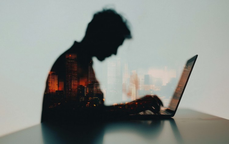 Silhouette of a person working on a laptop, superimposed with a cityscape at night.  The image is dark and moody, conveying a sense of late-night work or contemplation